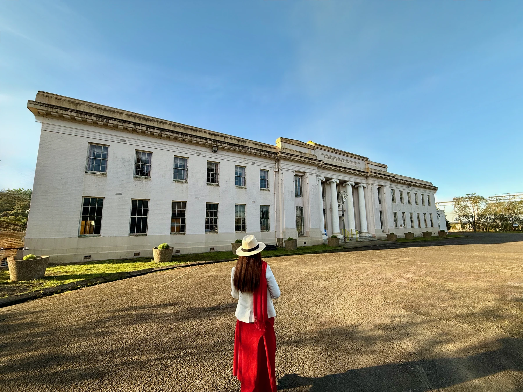 A person in a white jacket, red scarf and red trousers stands facing the white facade of the Yallourn Production Centre beneath a blue sky.