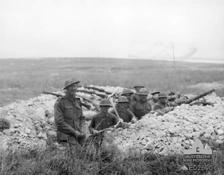 Australian and American soldiers stand and sit together in a shallow trench at Hamel, with open ground behind them.