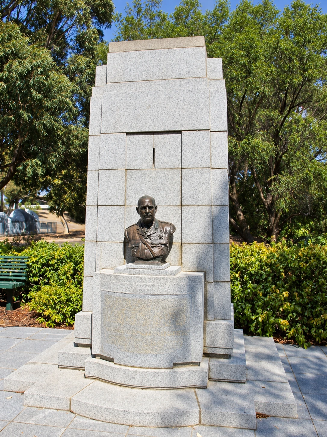 Restored colour portrait view showing the bronze bust of Sir John Monash against the pale stepped stone memorial, framed by trees and hedges.
