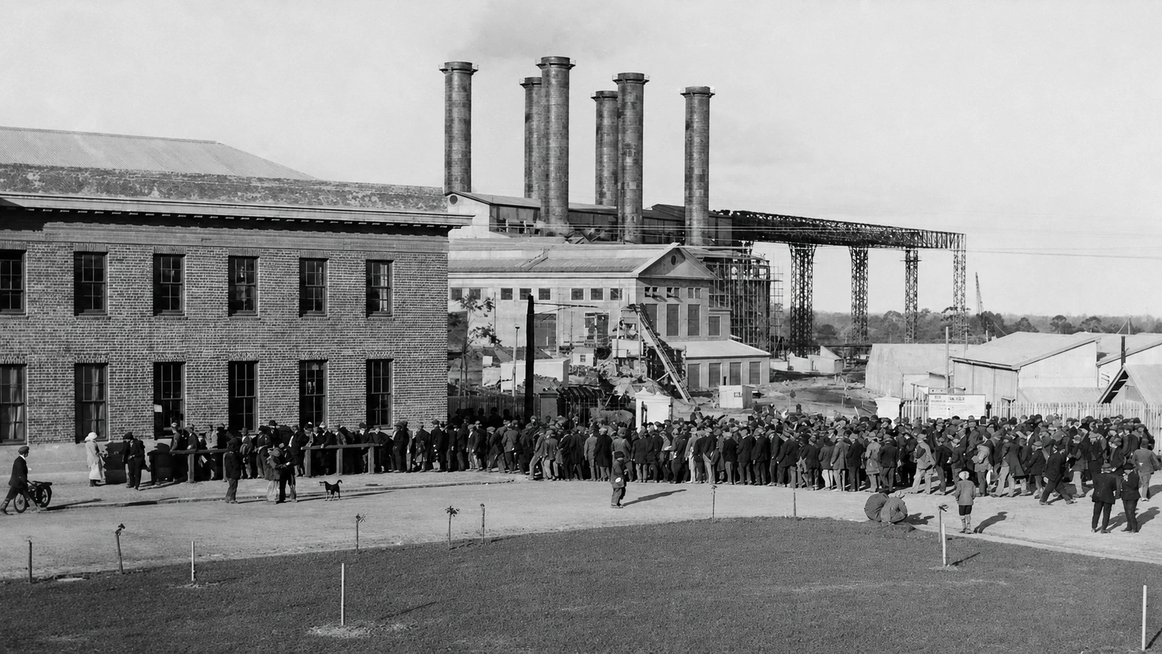 A long line of workers gathers outside the Yallourn power-station complex, with six chimneys and coal-handling structures behind them.