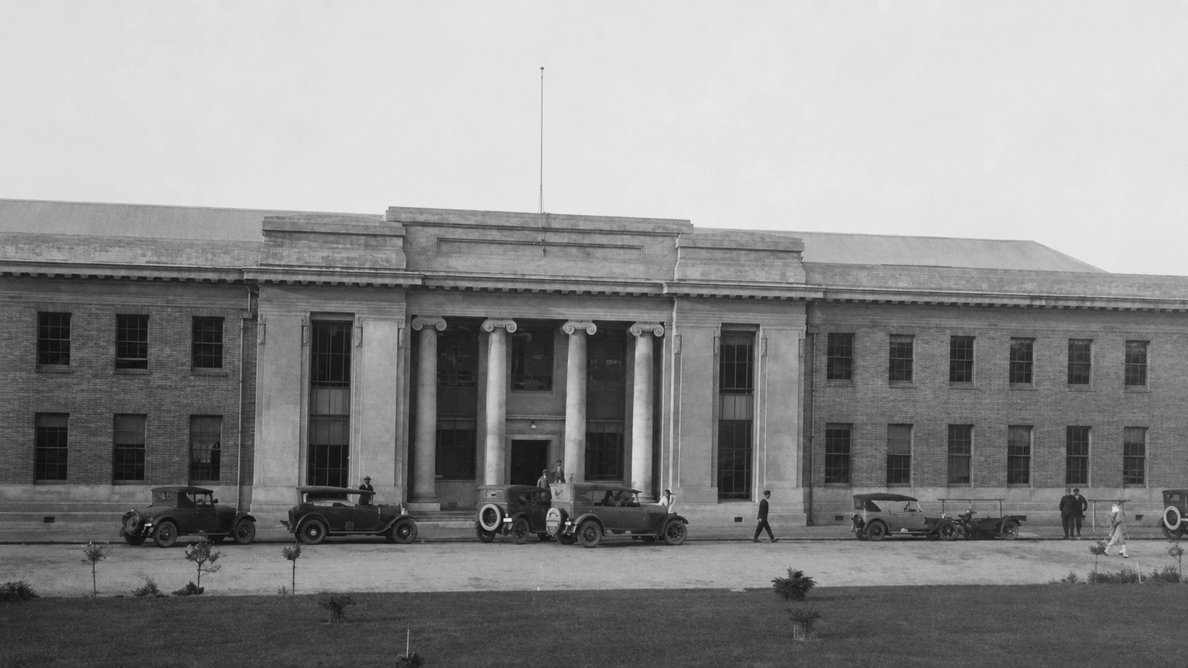 The columned front of the Yallourn Production Centre, with period motor cars parked along the road and several people near the entrance.
