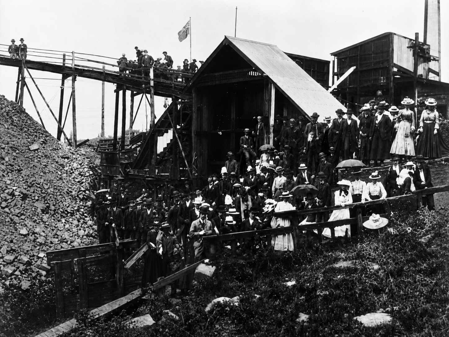 Restored black-and-white photograph of a large gathering of men, women and children among timber mine buildings and elevated coal-handling structures at Yallourn North.