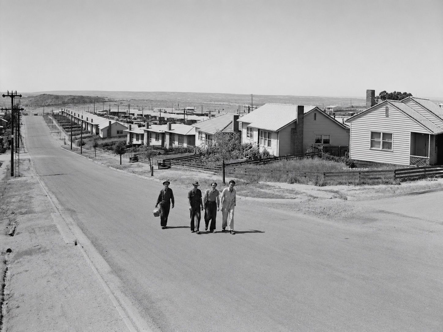 Restored black-and-white photograph of four workers walking up North Road in Yallourn North, with postwar houses lining the hillside behind them.