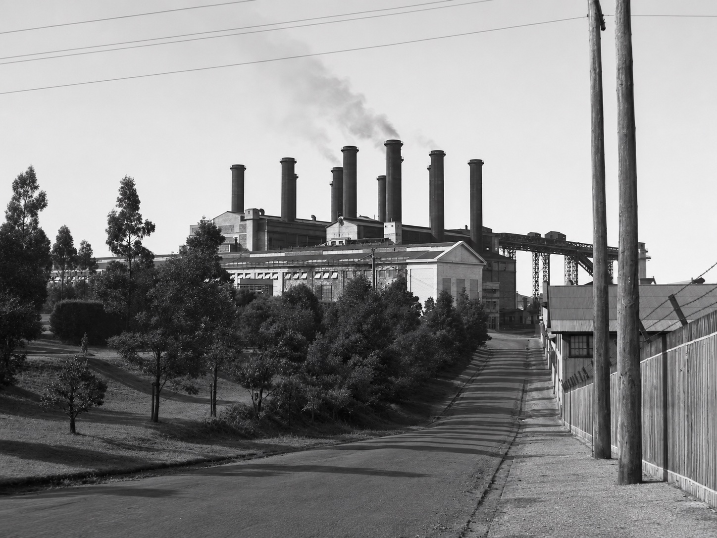 Restored black-and-white view along a road towards Yallourn Power Station, with gardens, industrial buildings, power lines and multiple tall chimneys.