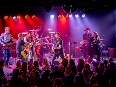 A band performing on a pub stage bathed in warm stage lighting, capturing the gritty rock atmosphere of a John Mellencamp tribute show.