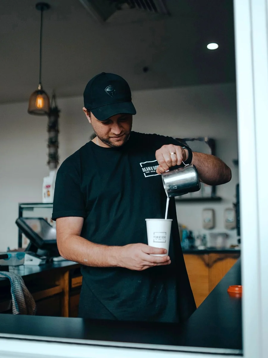Dean pours milk from a metal jug into a takeaway cup at the Bean n' Gone service window.