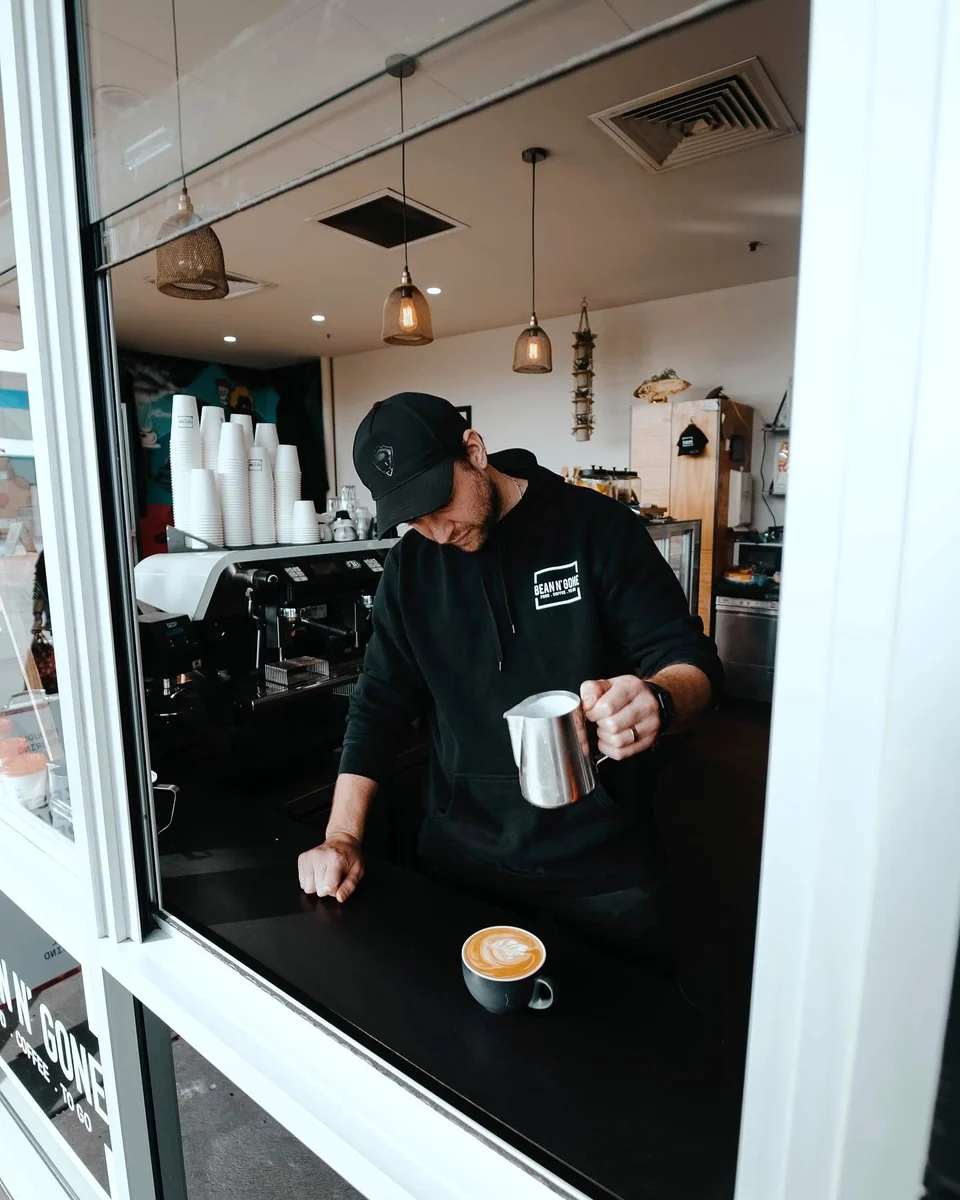 Dean works at the Bean n' Gone coffee window with a cup on the counter and a milk jug in hand.