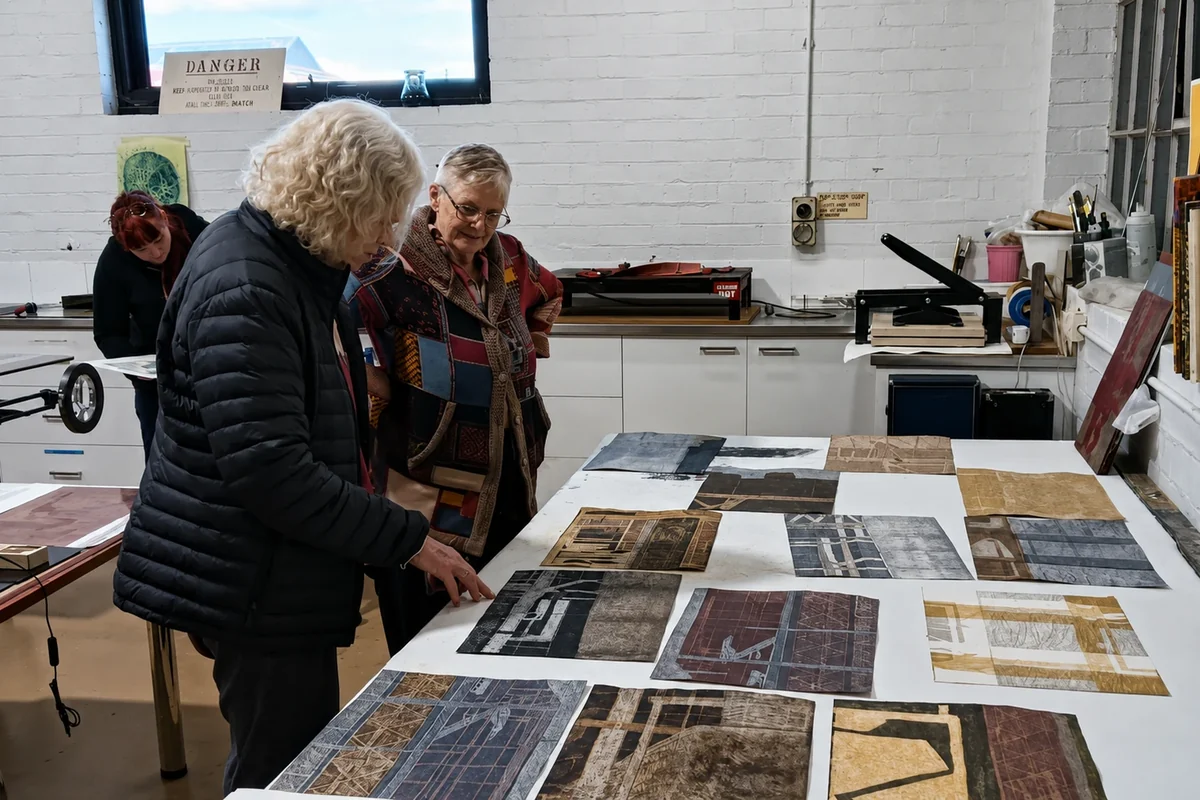 Three people examine a table covered with prints inside ARC Yinnar's white-brick printmaking room.