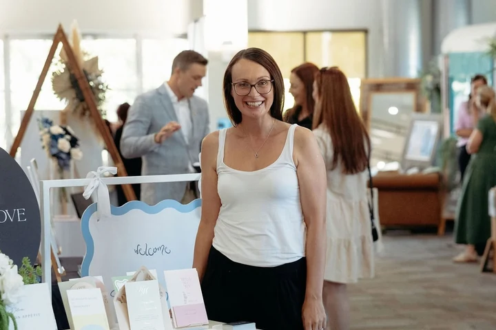 A smiling woman stands beside a wedding display during an indoor fair.