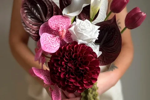 A person holds a sculptural bouquet of deep burgundy, pink and white flowers.