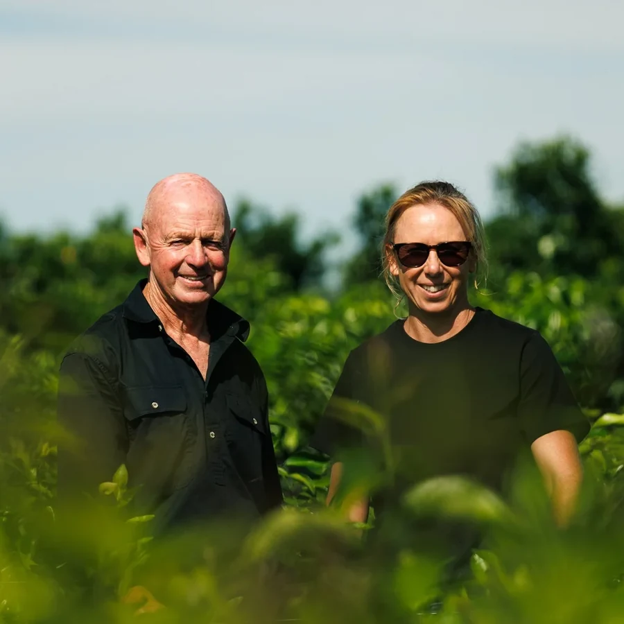 Two nursery growers in black shirts stand together among green trees.