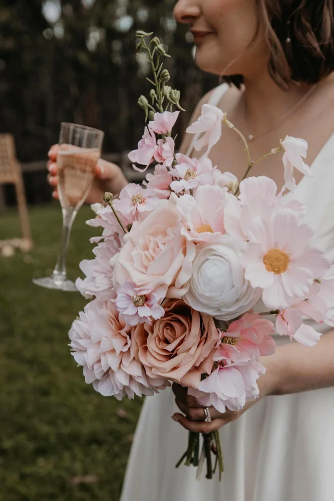 A person in white holds a large pale pink and cream bouquet beside a glass of sparkling drink.