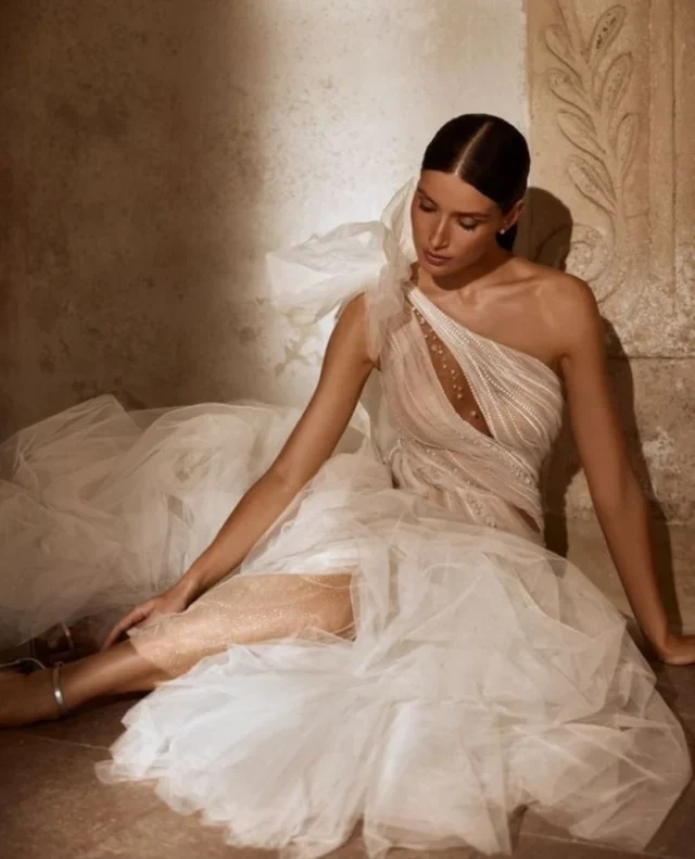 A seated model wears a layered tulle wedding gown with an asymmetric bodice.