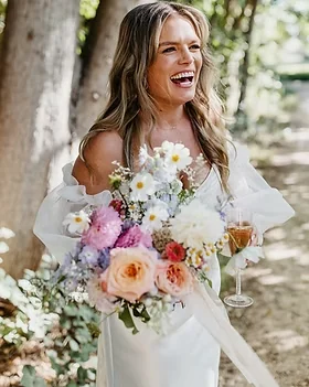 A bride holds a soft pink and white bouquet and a glass outdoors.