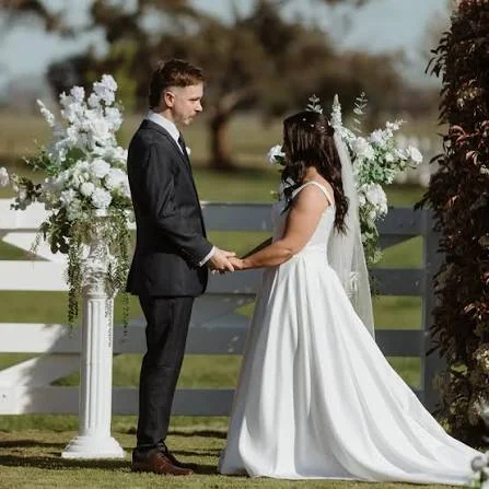 A bride and groom hold hands outdoors between tall white floral arrangements.