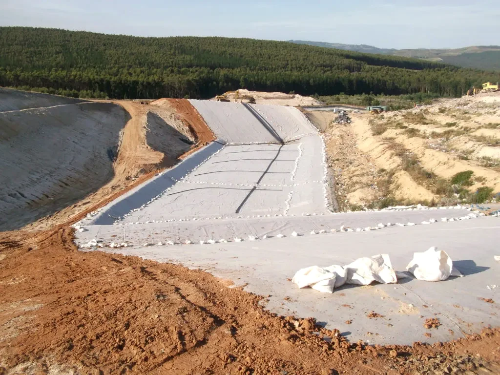 A large landfill cell lined with pale membrane beside forested hills.